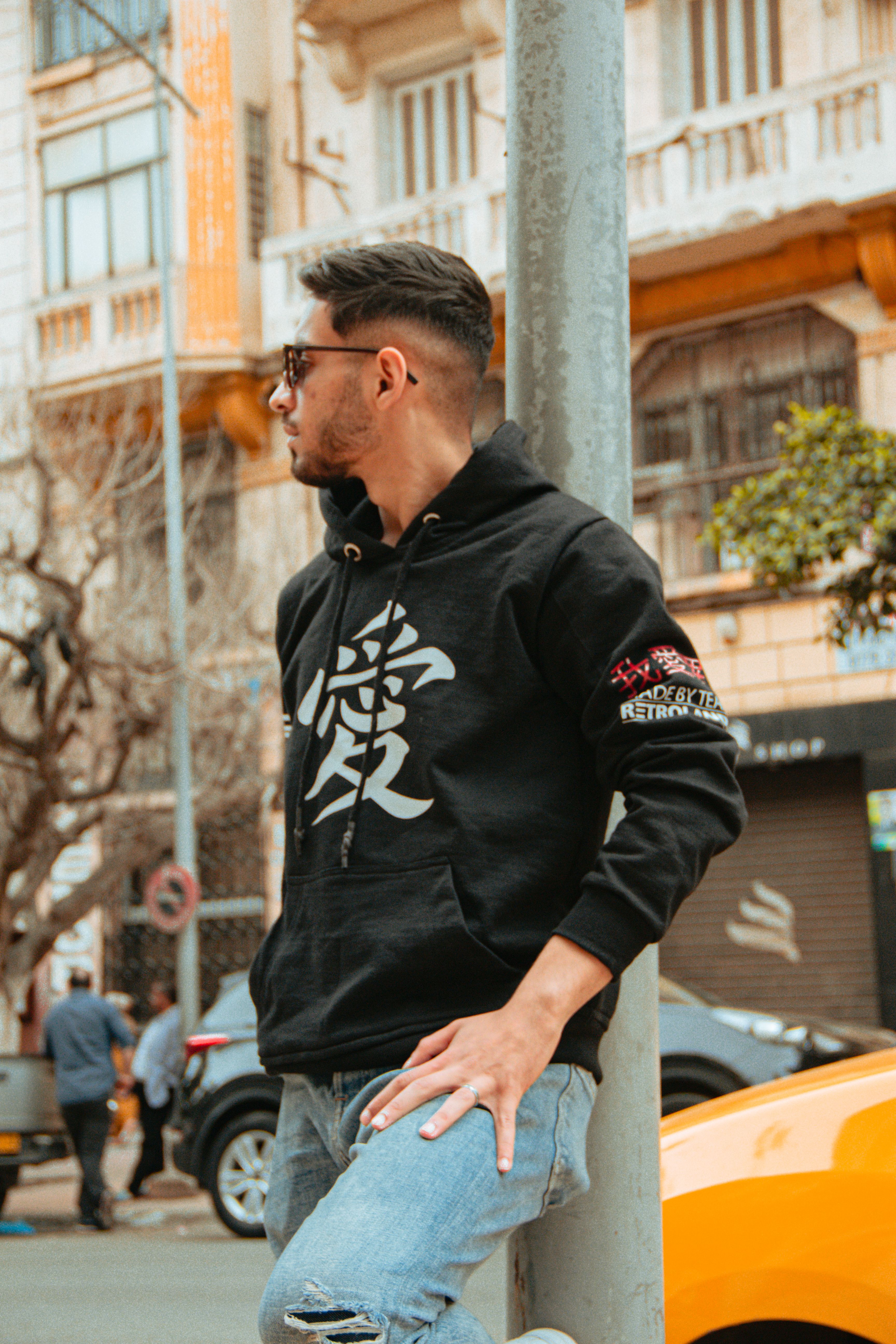 Young man in hoodie leaning on pole with urban background, wearing sunglasses.