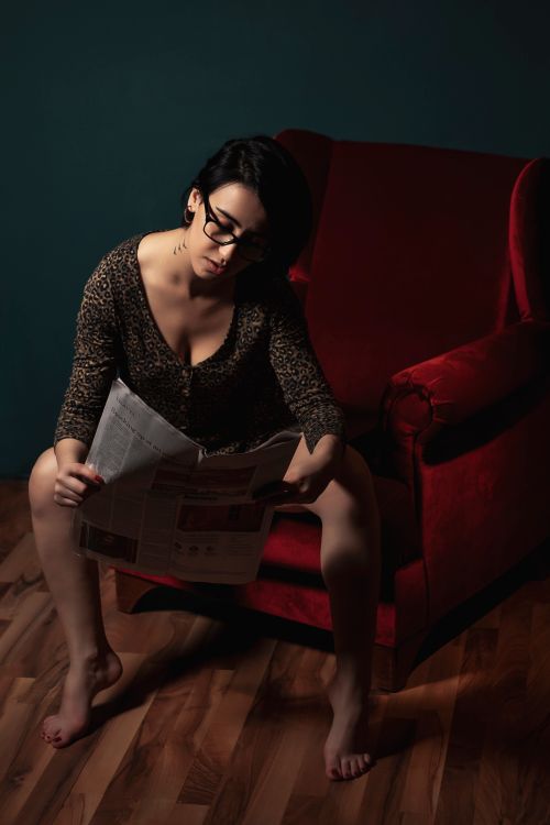 Free Adult woman in eyeglasses reads a newspaper barefoot while sitting on a red chair indoors. Stock Photo