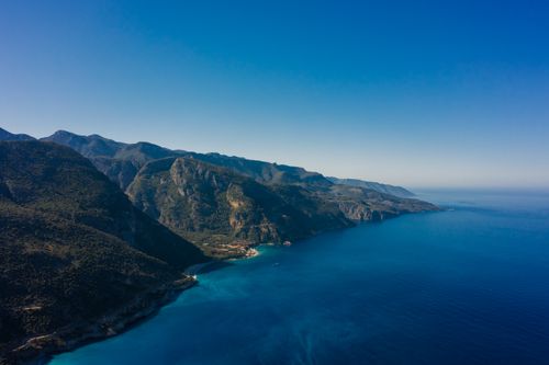 Free A breathtaking aerial shot of a rugged coastline with mountains and a vibrant blue sea under a clear sky. Stock Photo
