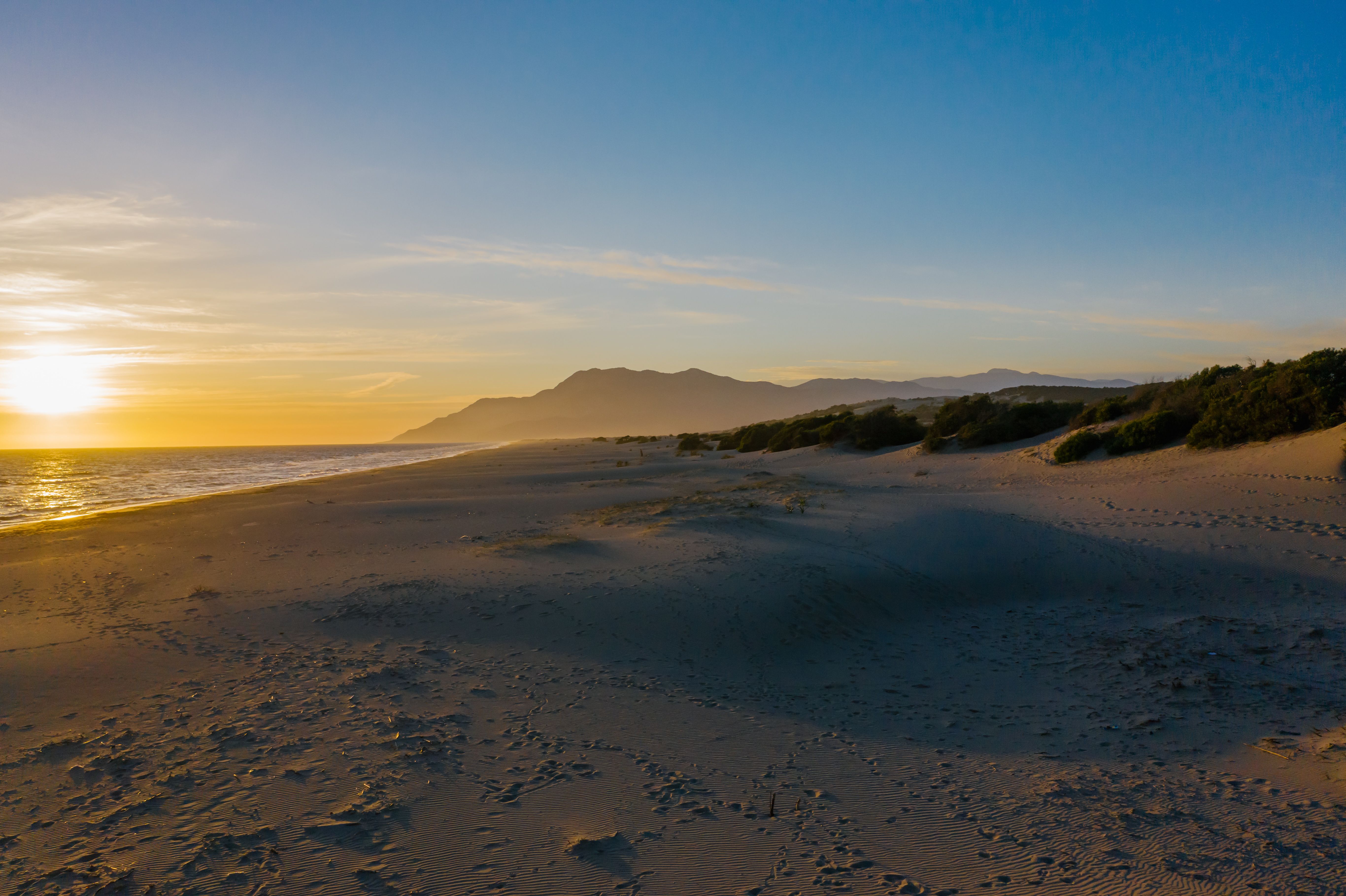 View of a Beach, Mountains in Distance and Sun Setting above the Sea ...