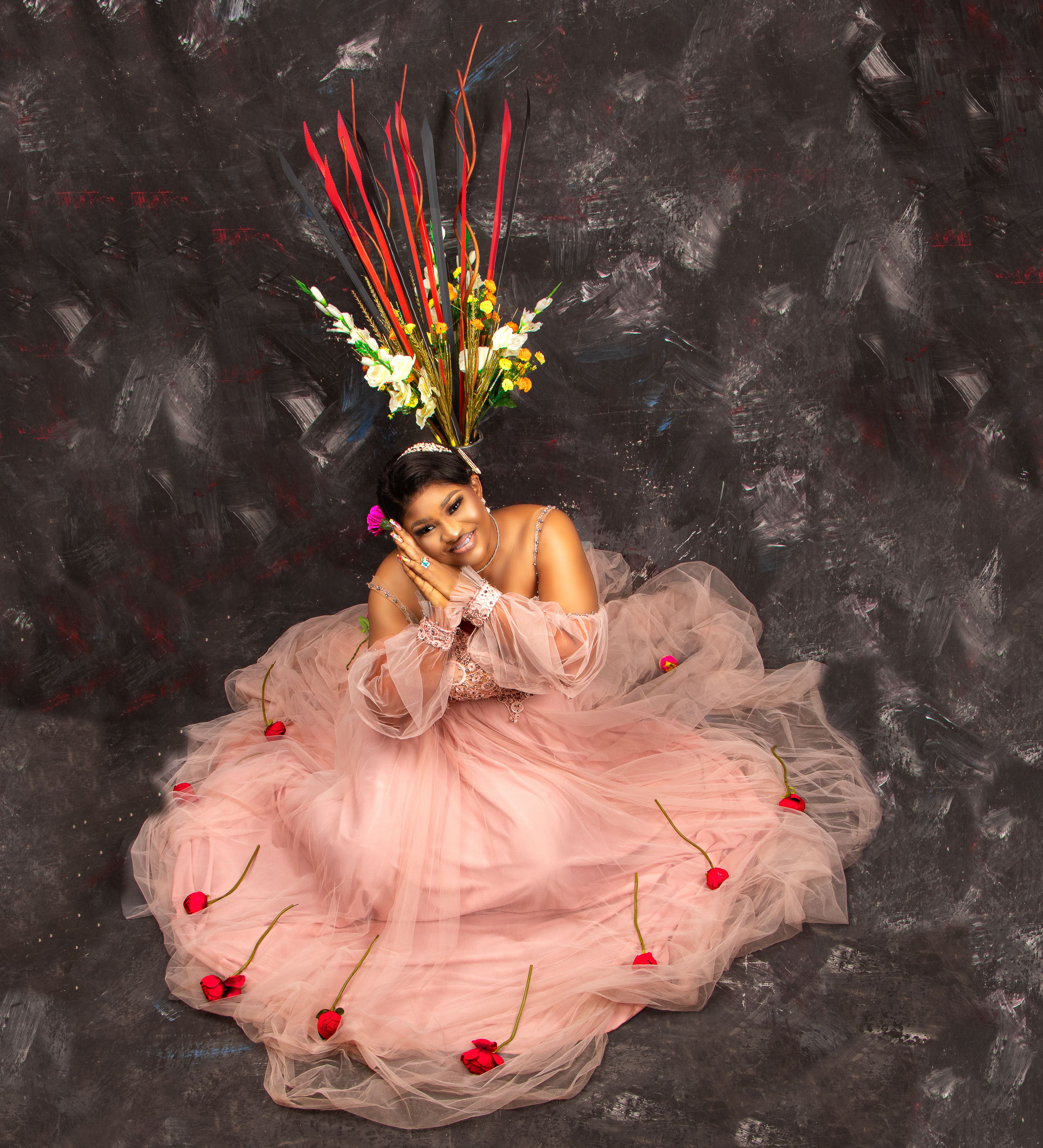 Cheerful African American female model with flowers in hair wearing stylish long pink dress looking at camera on gray background in studio