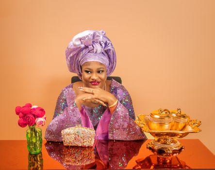 Happy adult African American female with makeup in silk turban and elegant dress covered with sequins sitting at table with hands at chin and looking away happily
