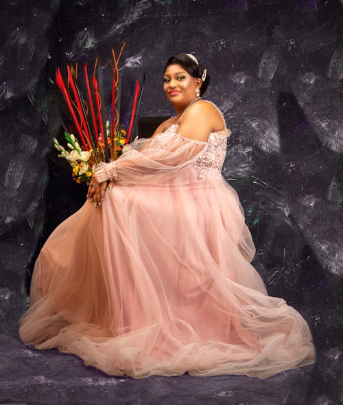Side view of happy elegant ethnic lady with dark hair in maxi pink chiffon dress sittinng on chair in studio and looking at camera