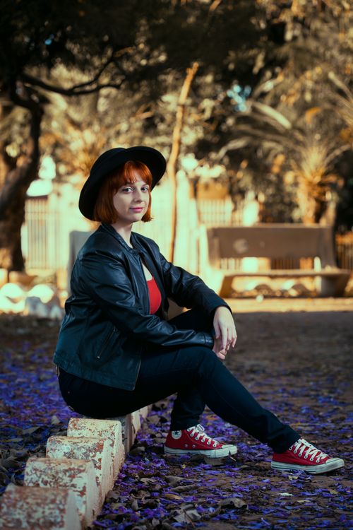 Free Woman with hat and leather jacket sitting on park ledge. Autumn vibes, stylish fashion. Stock Photo