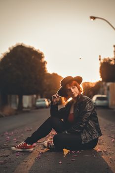 Fashionable woman wearing a black leather jacket and hat, seated on a street at sunset.