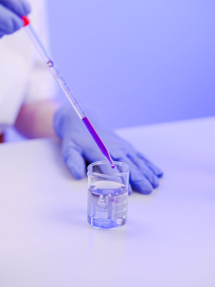 Close-Up View Of A Person Pipetting A Chemical Into A Small Beaker