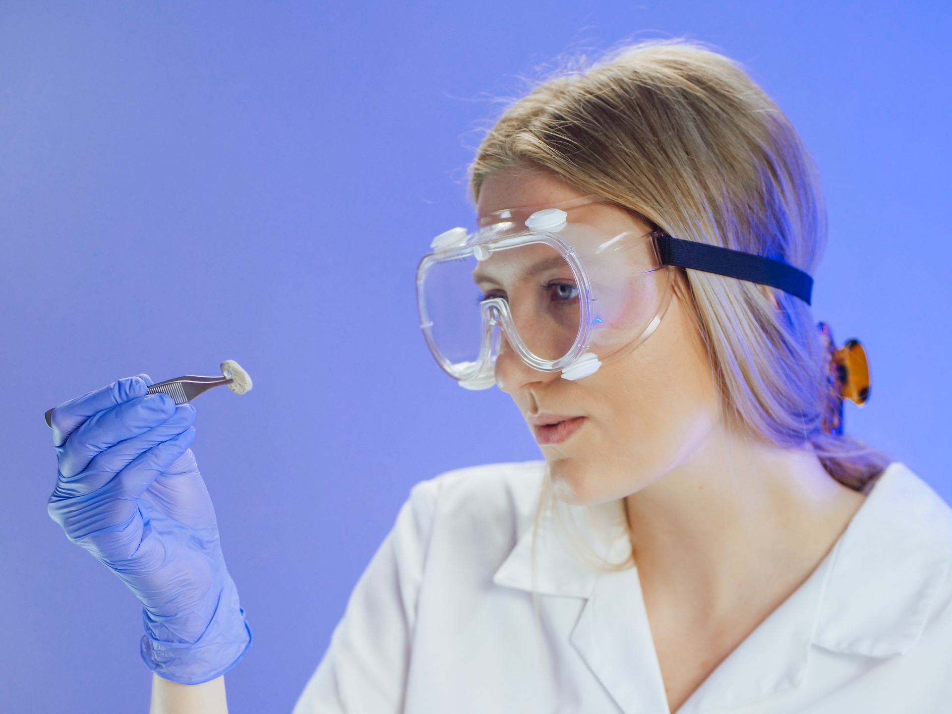 A female scientist examining a sample with forceps in a laboratory, wearing safety goggles and gloves.