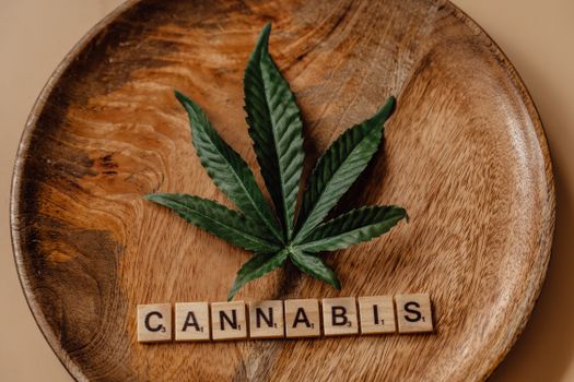 Top view of a cannabis leaf on a wooden plate with Scrabble tiles spelling 'cannabis'.