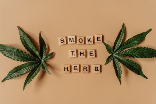 Free Flat lay of cannabis leaves and scrabble tiles spelling 'Smoke the Herb' on a brown background. Stock Photo