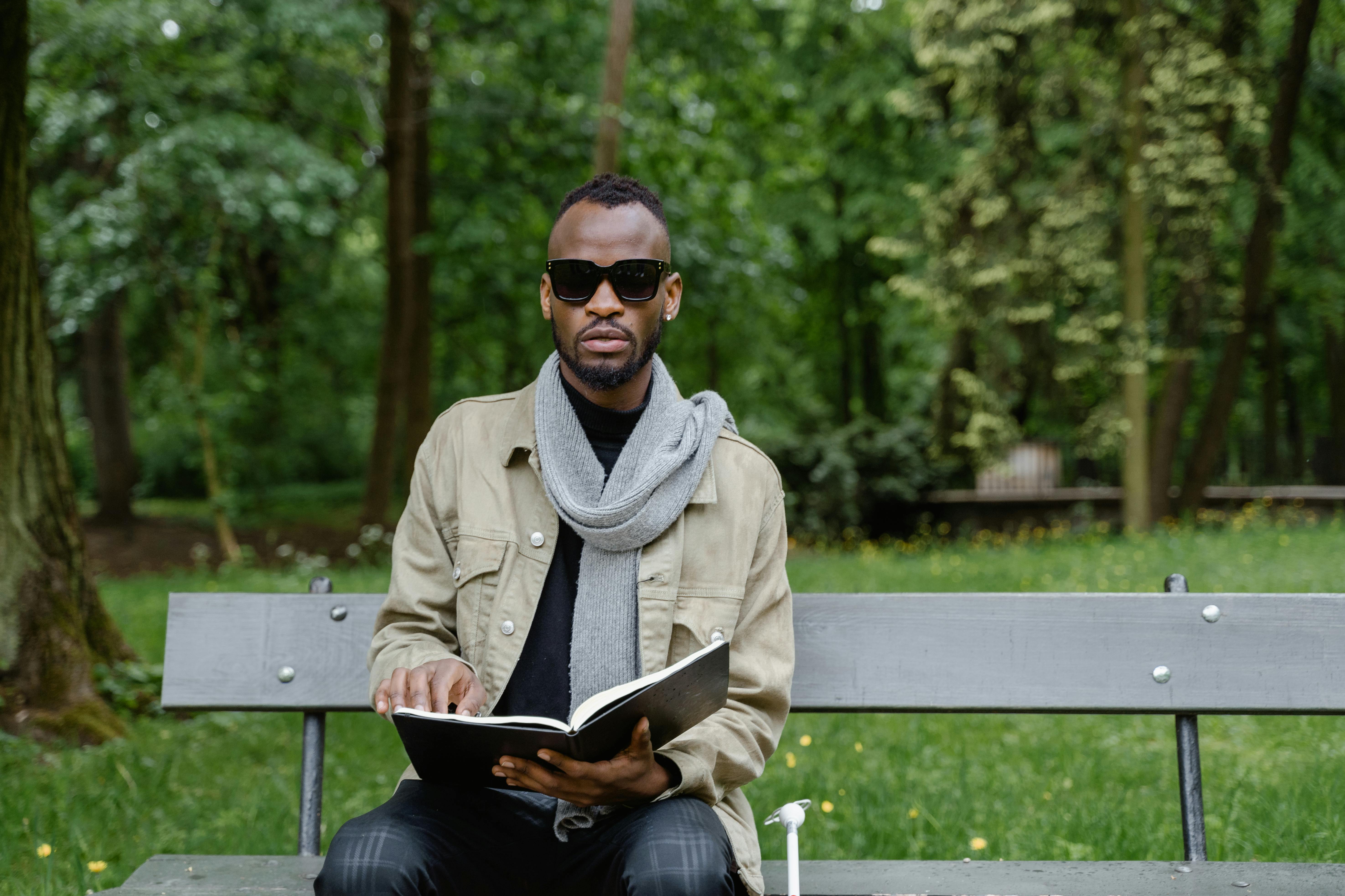 African American man using Braille book seated outdoors on a park bench wearing sunglasses and a scarf.