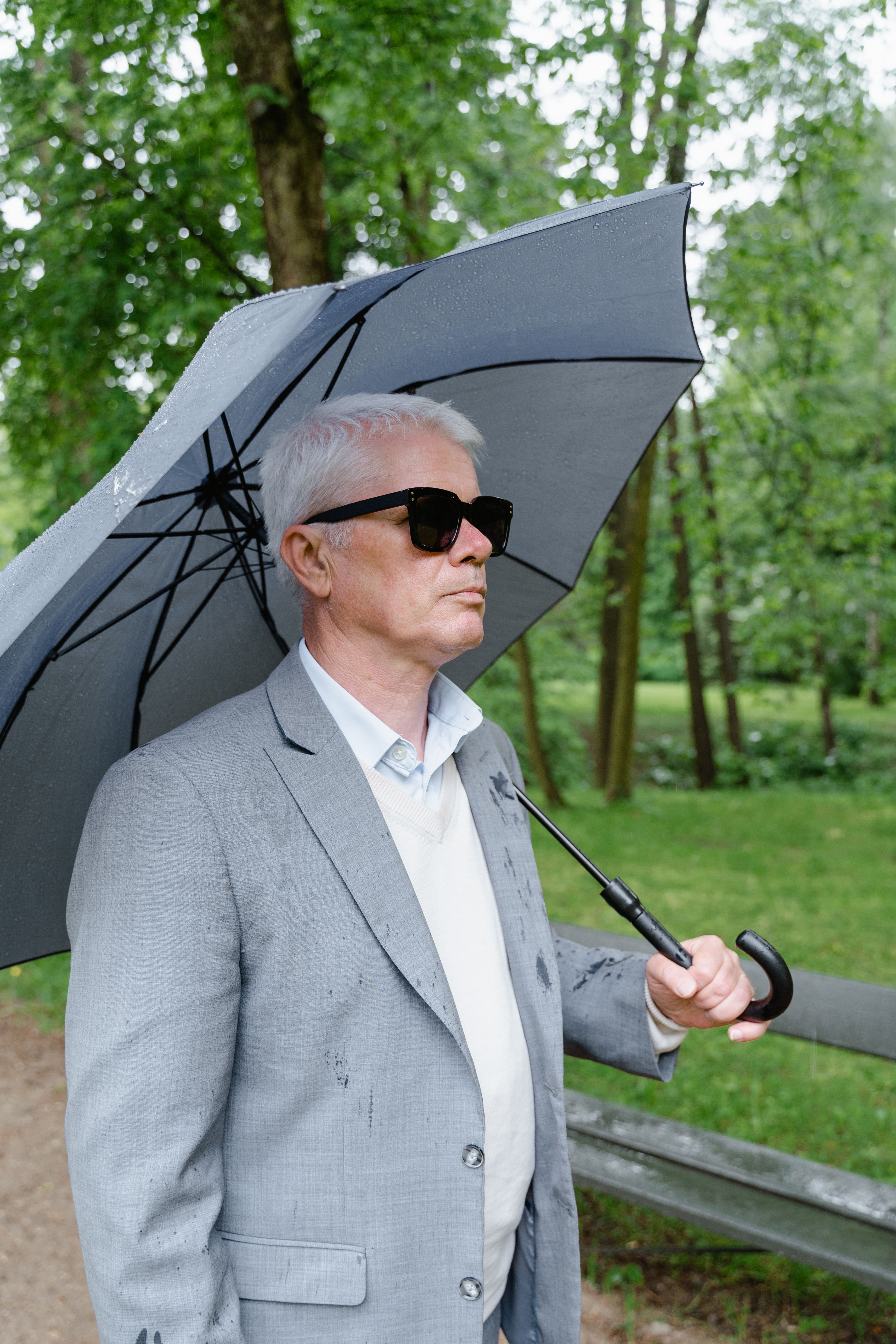 Senior man in a gray suit holding an umbrella in a park, exuding style and elegance.