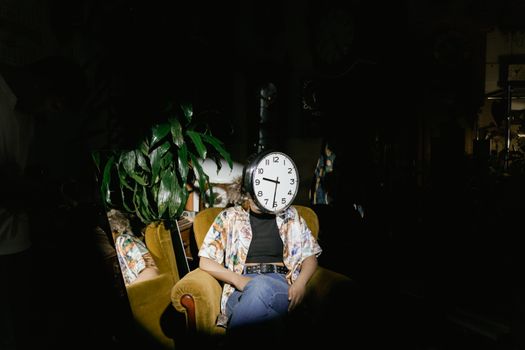 A unique portrait of a person with a clock as a head, seated in a dimly lit room with houseplants.
