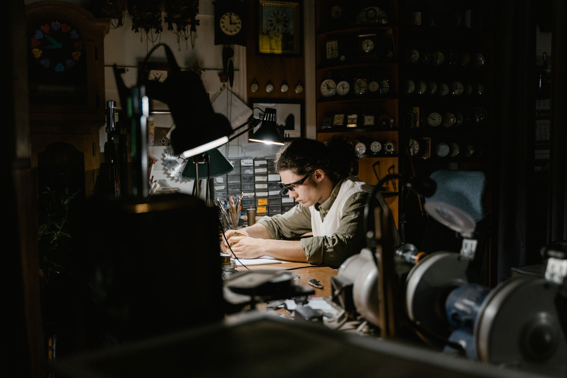 A skilled watchmaker concentrating on repairing clocks in a dimly lit workshop surrounded by tools.