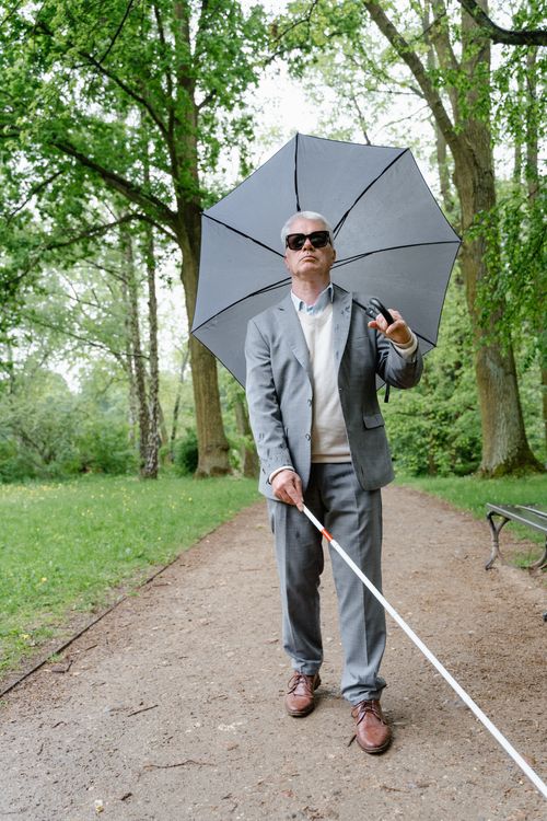 Free Elderly man with sunglasses and cane on a park path using an umbrella. Stock Photo