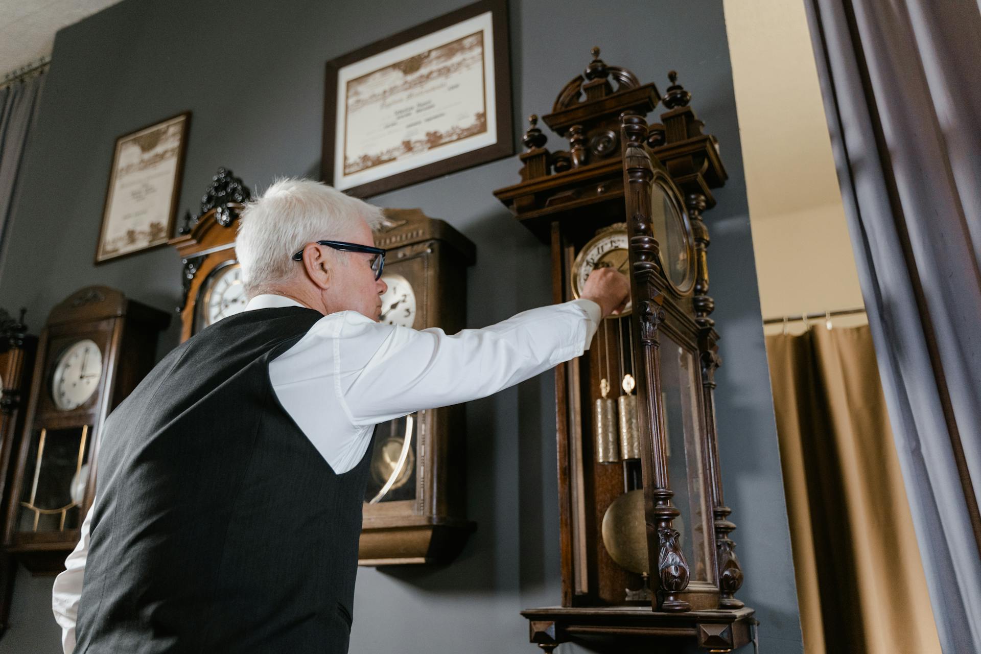 An elderly man adjusts an antique grandfather clock inside a vintage clock shop.