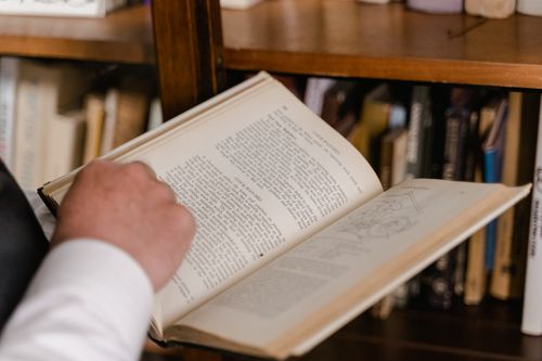 Free Focused image of a hand holding open a book against a bookshelf, emphasizing reading and knowledge. Stock Photo