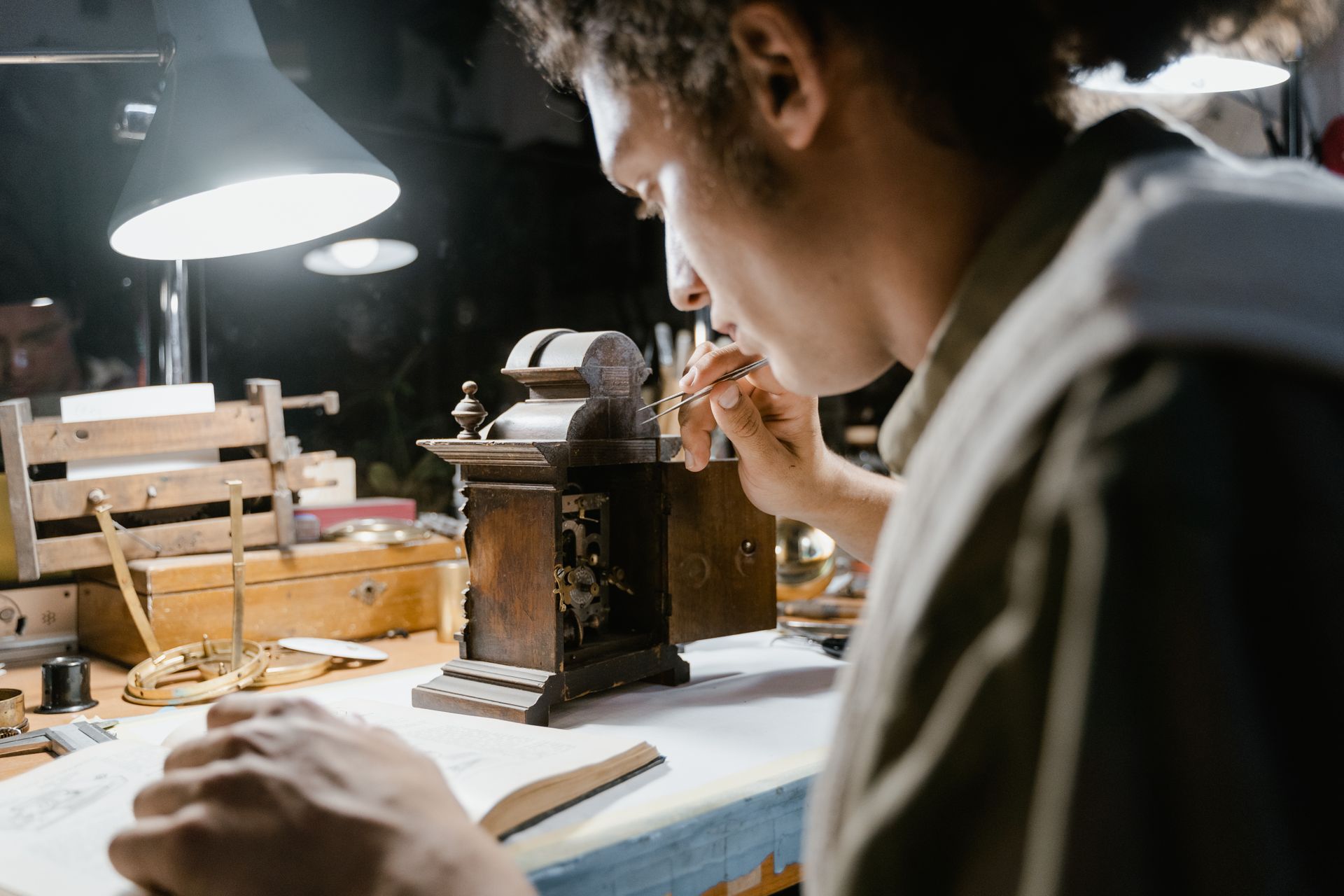 A focused artisan carefully restores an antique clock with precision tools under lamp lighting.