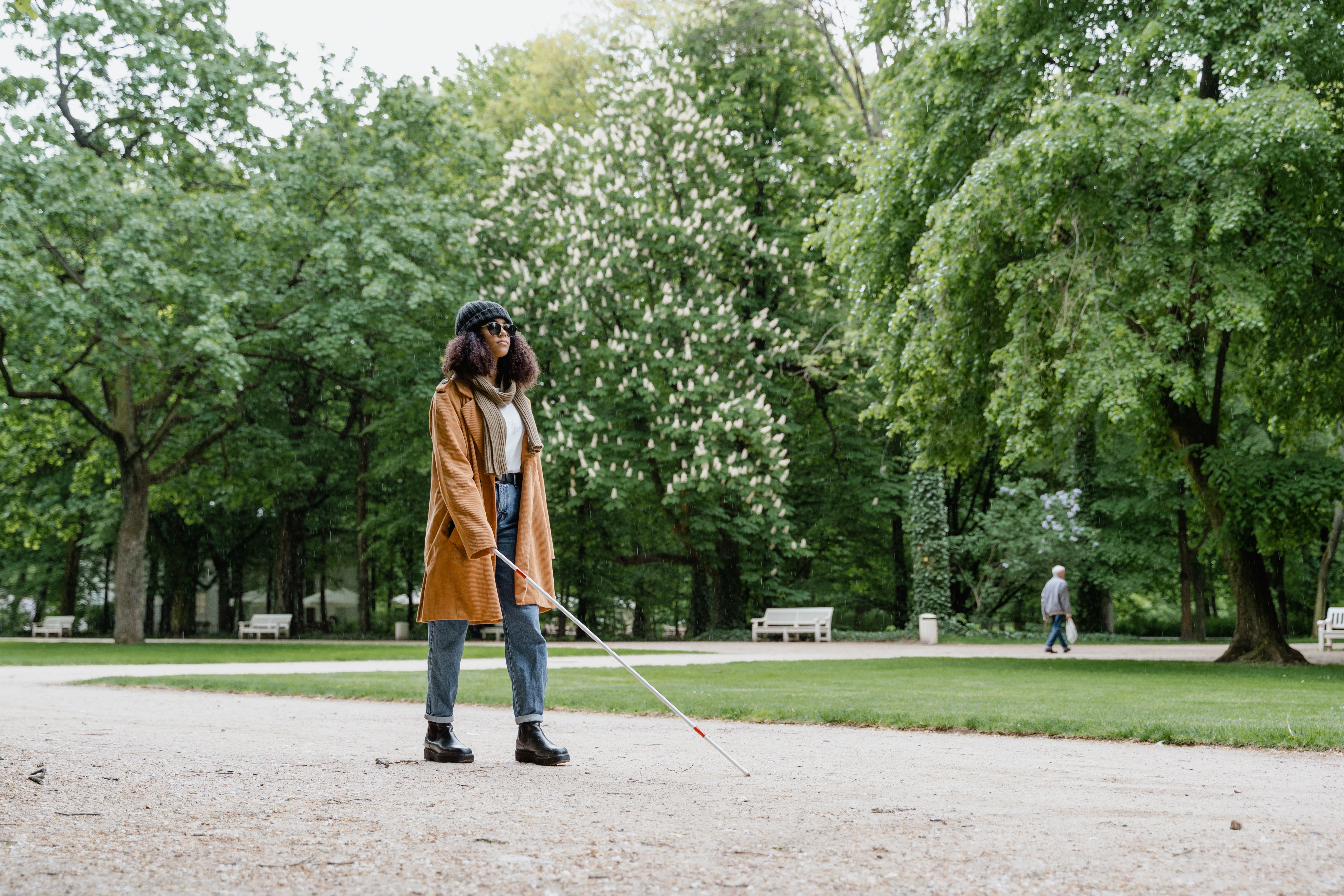 A blind woman using a white cane walks through a lush green park on a sunny day.