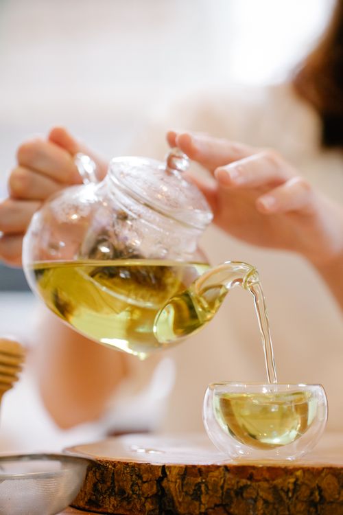 Free A close-up of green tea being poured from a clear glass teapot into a matching teacup on a wooden surface. Stock Photo