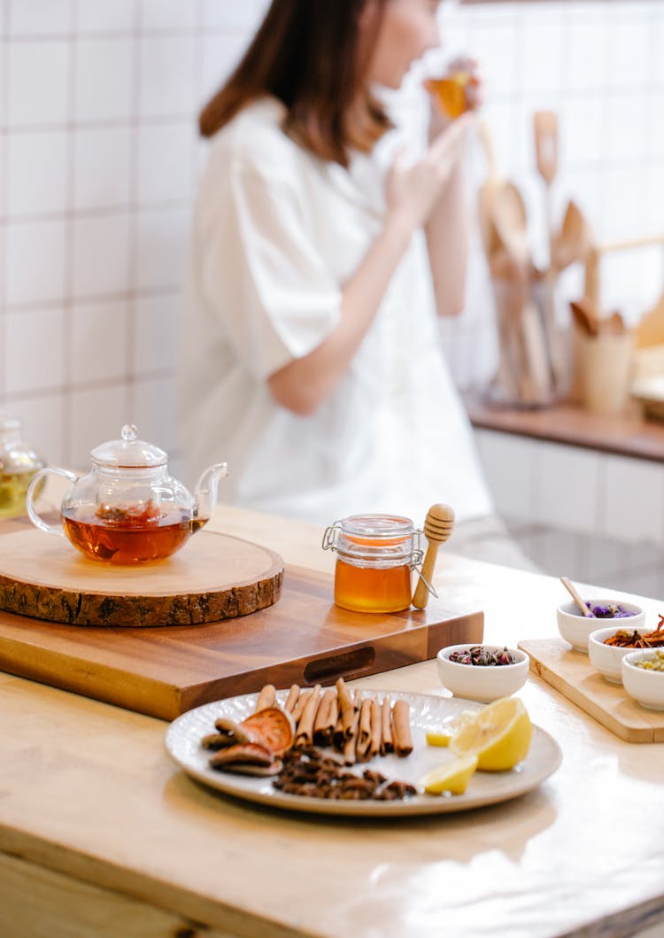 Woman Standing Behind A Counter In The Kitchen With Teapot, Honey And A Plate With Spices 