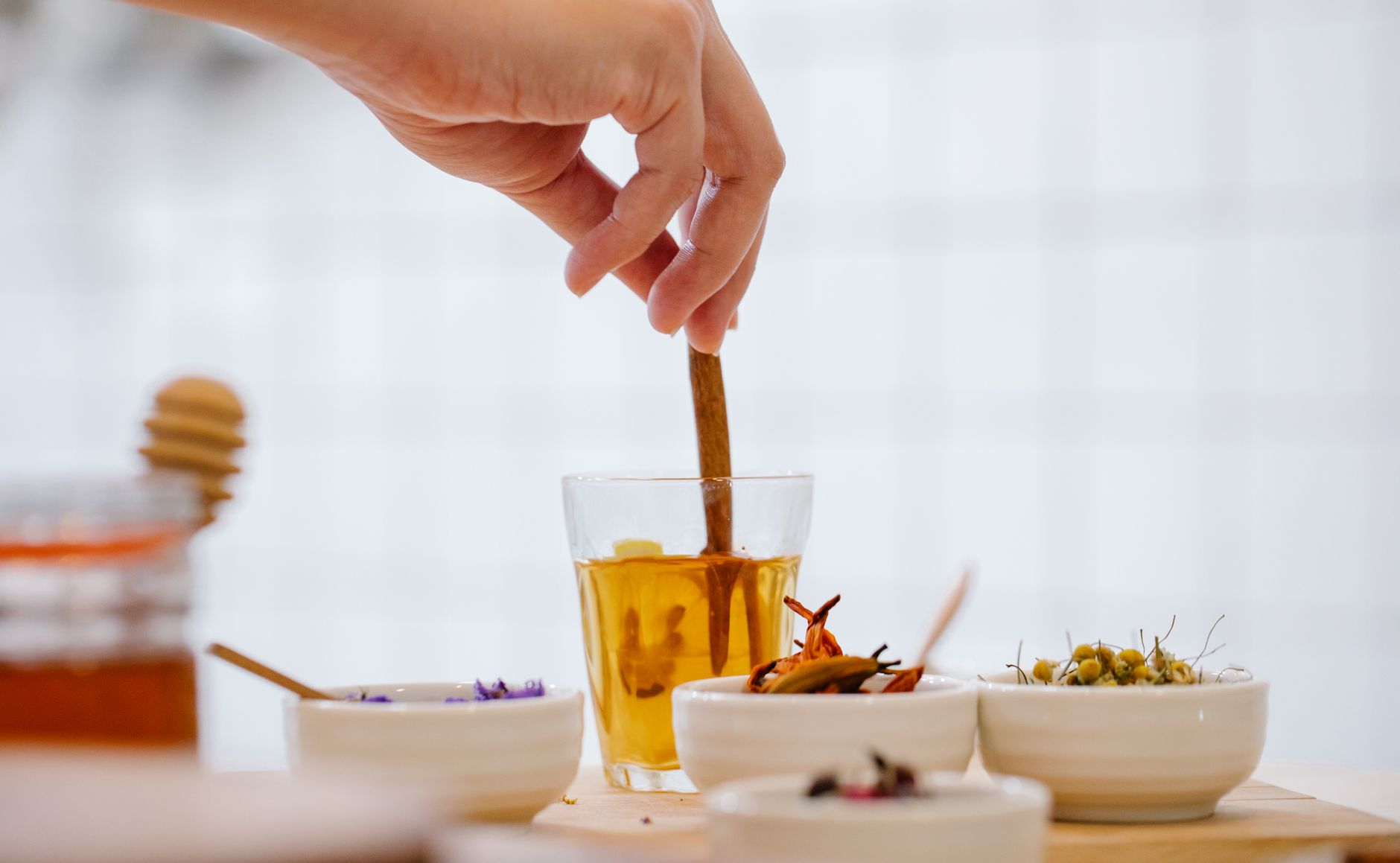 A hand stirs herbal tea in a glass with bowls of spices and herbs on a kitchen counter.