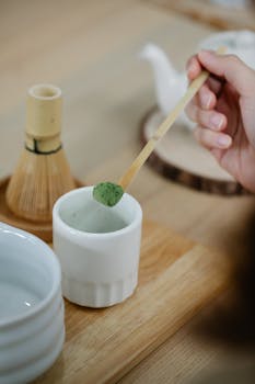 Hand holding bamboo spoon with matcha over traditional tea set, indoors on wooden table.