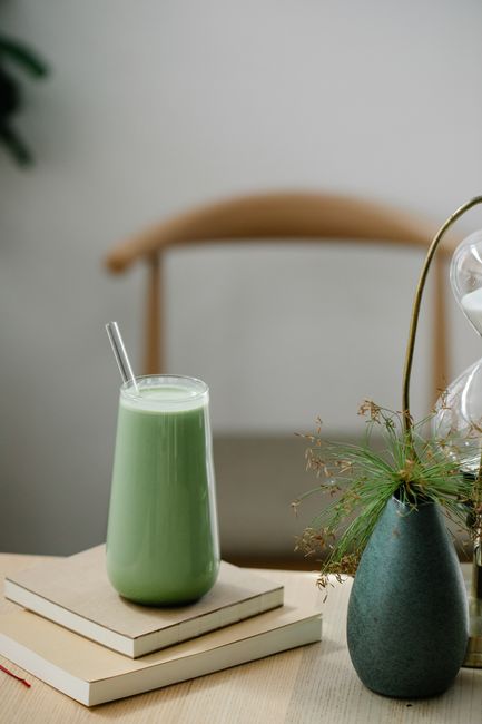 A chilled matcha latte with a straw beside books and a decorative vase on a table.