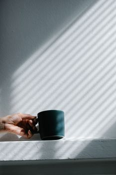 Minimalist composition of a black mug held by a hand with striped shadows on a white wall.