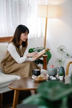 A woman sitting on a couch, reading a book in a cozy, well-lit room, enjoying relaxation.