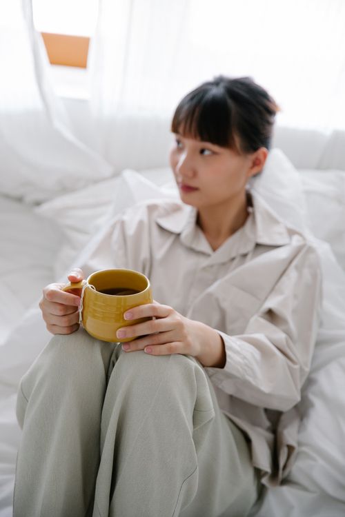 Free Asian woman in pajamas holding a tea mug, relaxed in bed interior. Stock Photo