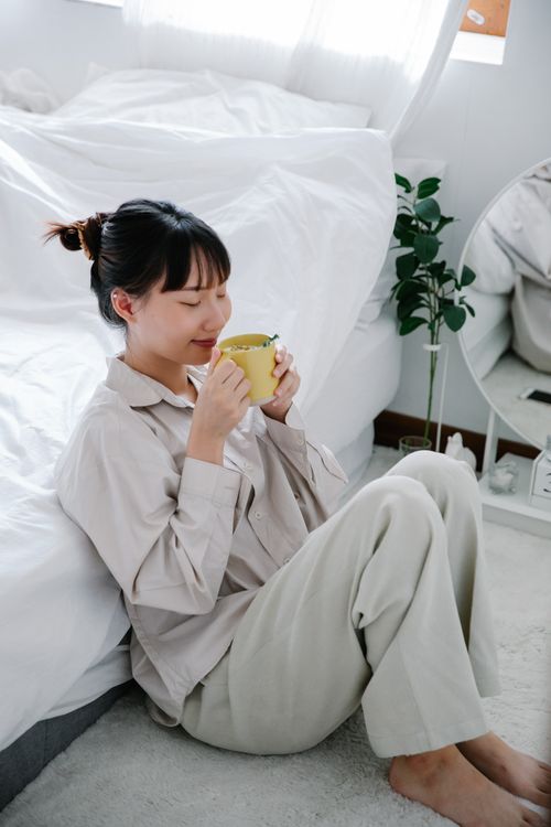 Free East Asian woman sipping tea, seated comfortably in a bright, cozy bedroom. Stock Photo