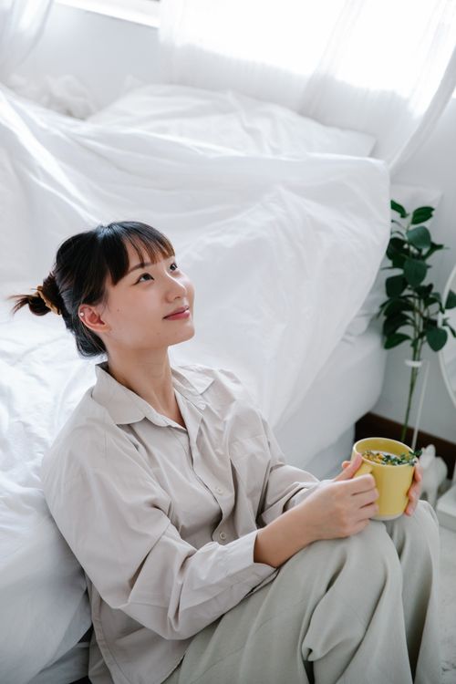 Free Asian woman enjoying a peaceful morning with tea in a bright bedroom, radiating tranquility. Stock Photo