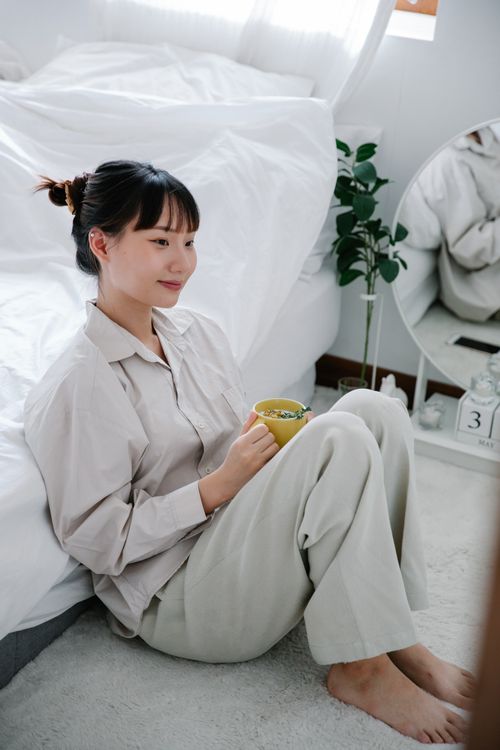 Free Woman sitting by her bed, enjoying a cup of tea in a bright, minimalist bedroom. Stock Photo