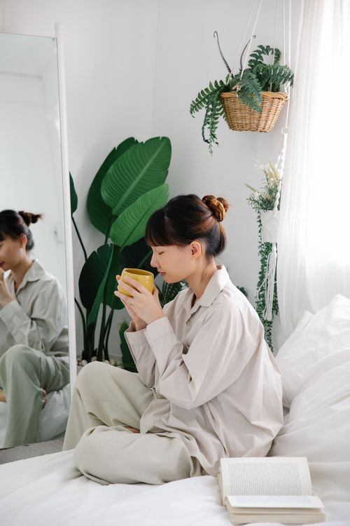 Free Woman sitting on bed with a mug, surrounded by lush indoor plants and open book. Stock Photo