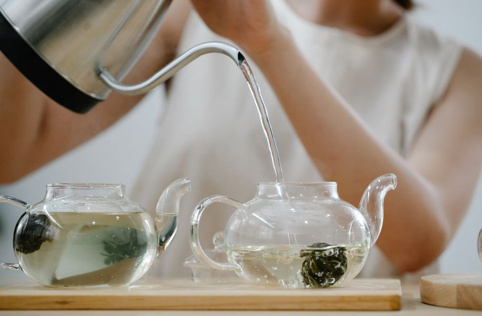 A woman pours hot water into glass teapots with blooming tea indoors.