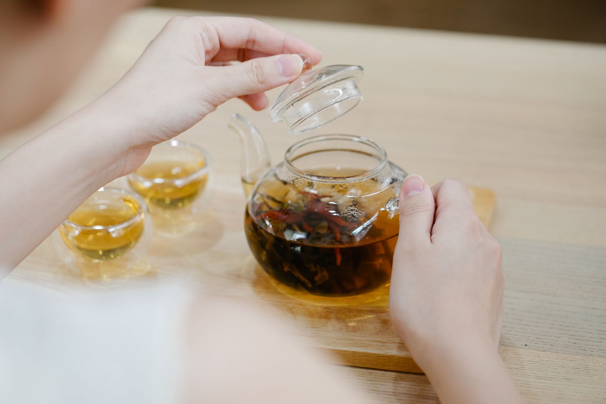 Hands lifting the lid of a glass teapot with herbal tea and two teacups nearby.