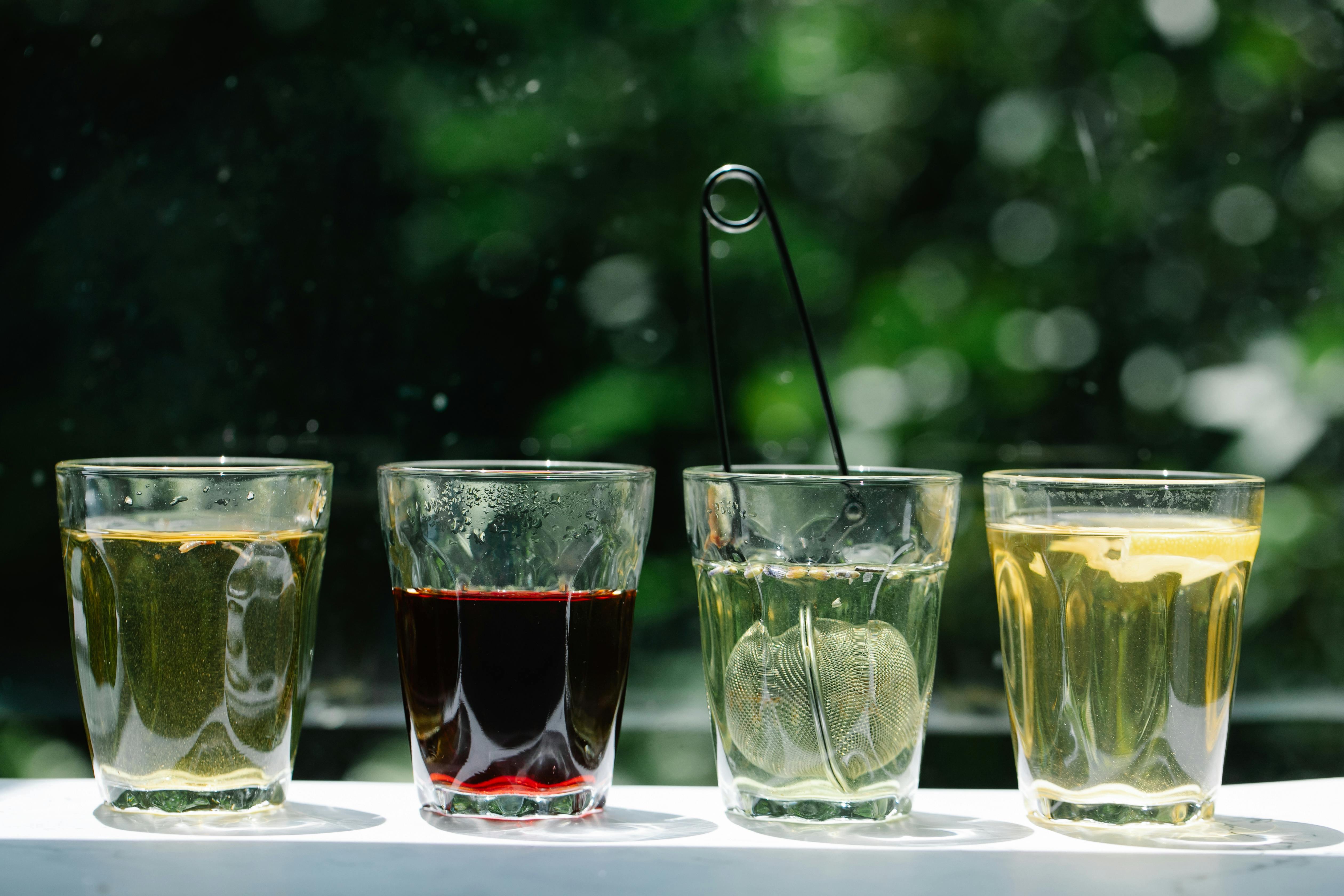 Four glasses of varying herbal teas with a strainer, set against a lush green background.