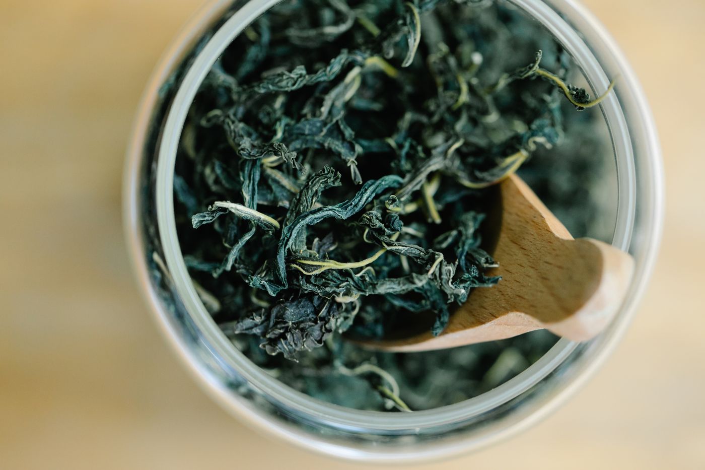 Curled dry green tea leaves in a glass jar with a wooden scoop