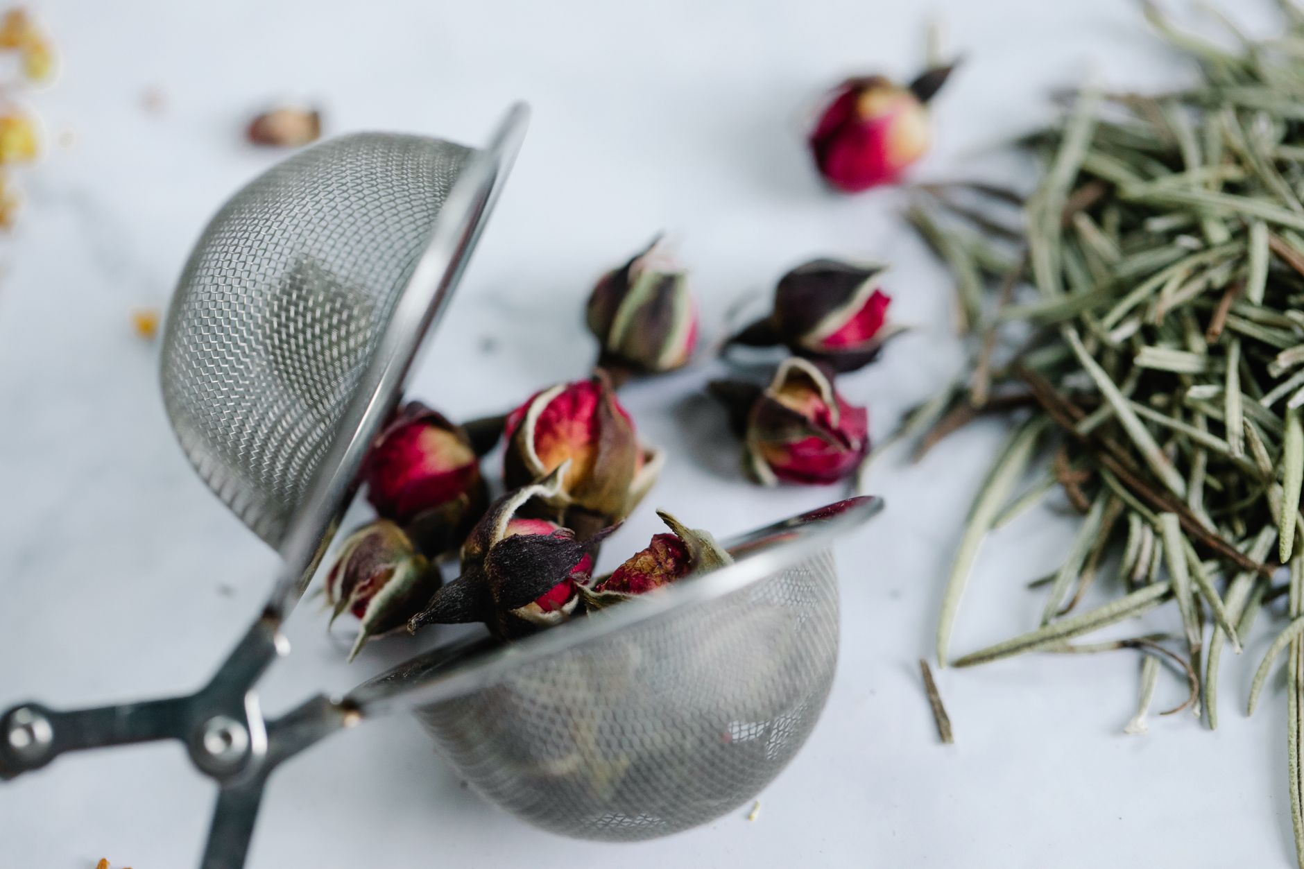 Rosemary Tea Recipe Homemade: A Fresh Brew for Wellness, Focus, and Flavor 2 Close-up of rosebuds and herbs in a stainless steel tea strainer on a white surface, highlighting rosemary tea recipe homemade ingredients.