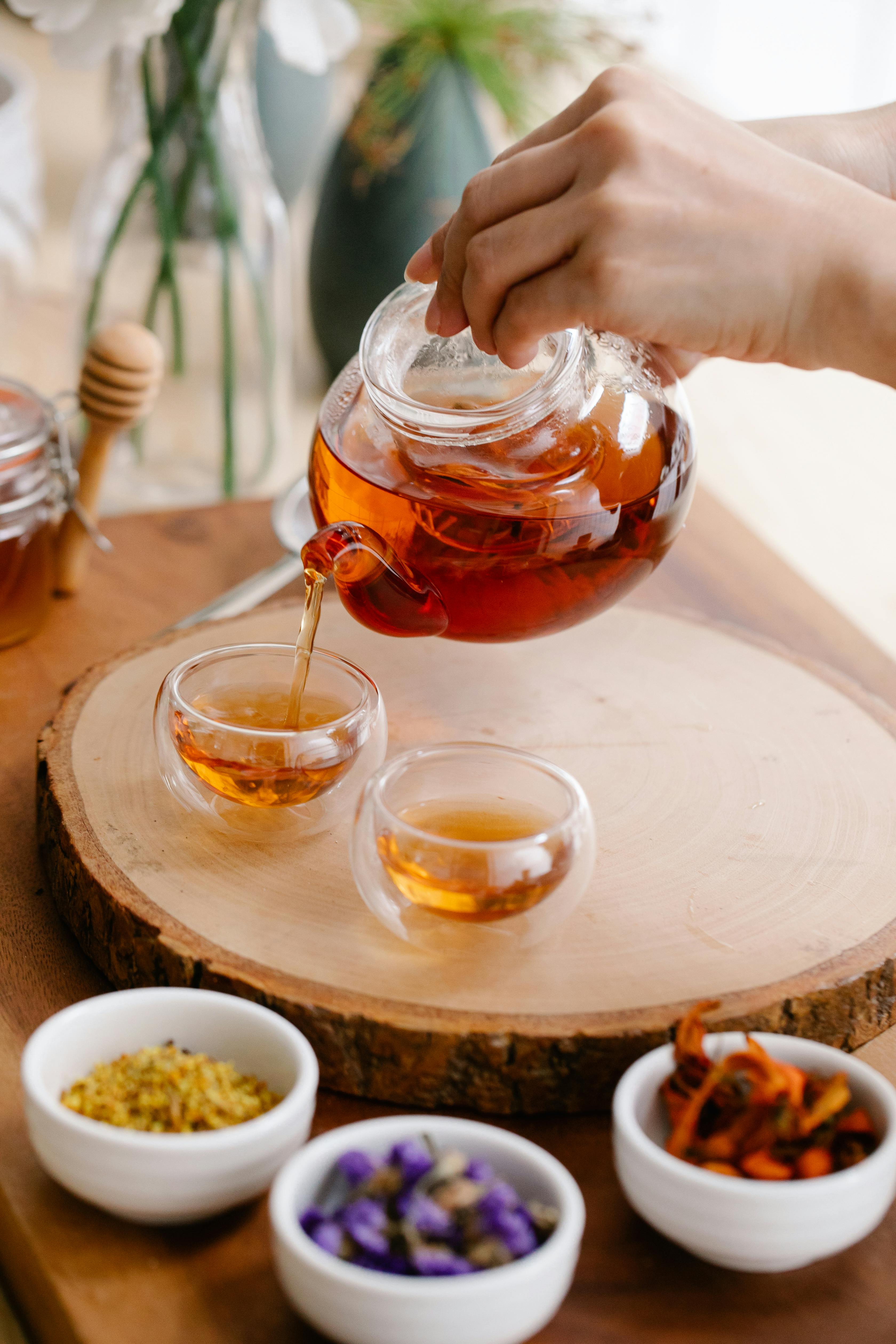 A person pouring hot Crowberry tea from a teapot into a glass cup, with a serene, natural background.