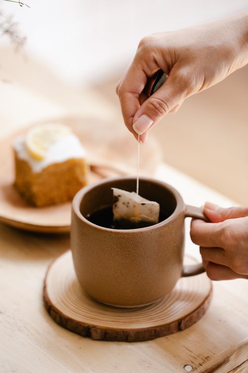 Free A hand dips a teabag into a brown mug of tea, next to a piece of lemon cake indoors. Stock Photo