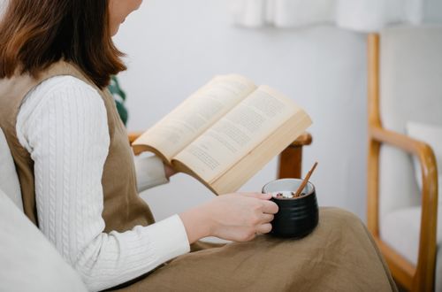 Free Woman sitting in an armchair reading a book and holding a cup of tea, in a peaceful indoor setting. Stock Photo