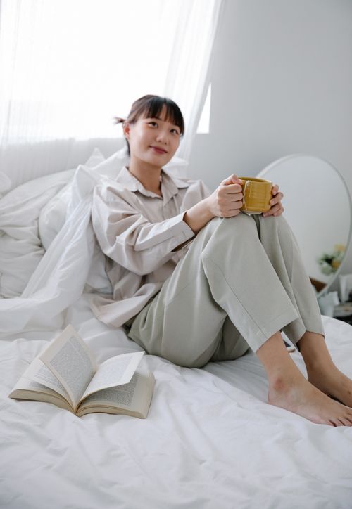 Free Asian woman enjoying a calm morning with a book and coffee in her comfortable bedroom setting. Stock Photo