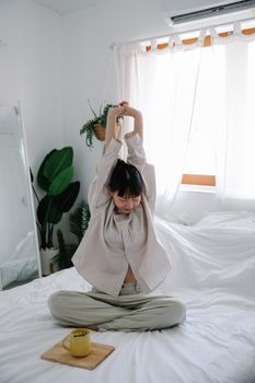 Woman in pajamas stretching on a bed in a sunlit bedroom with plants.