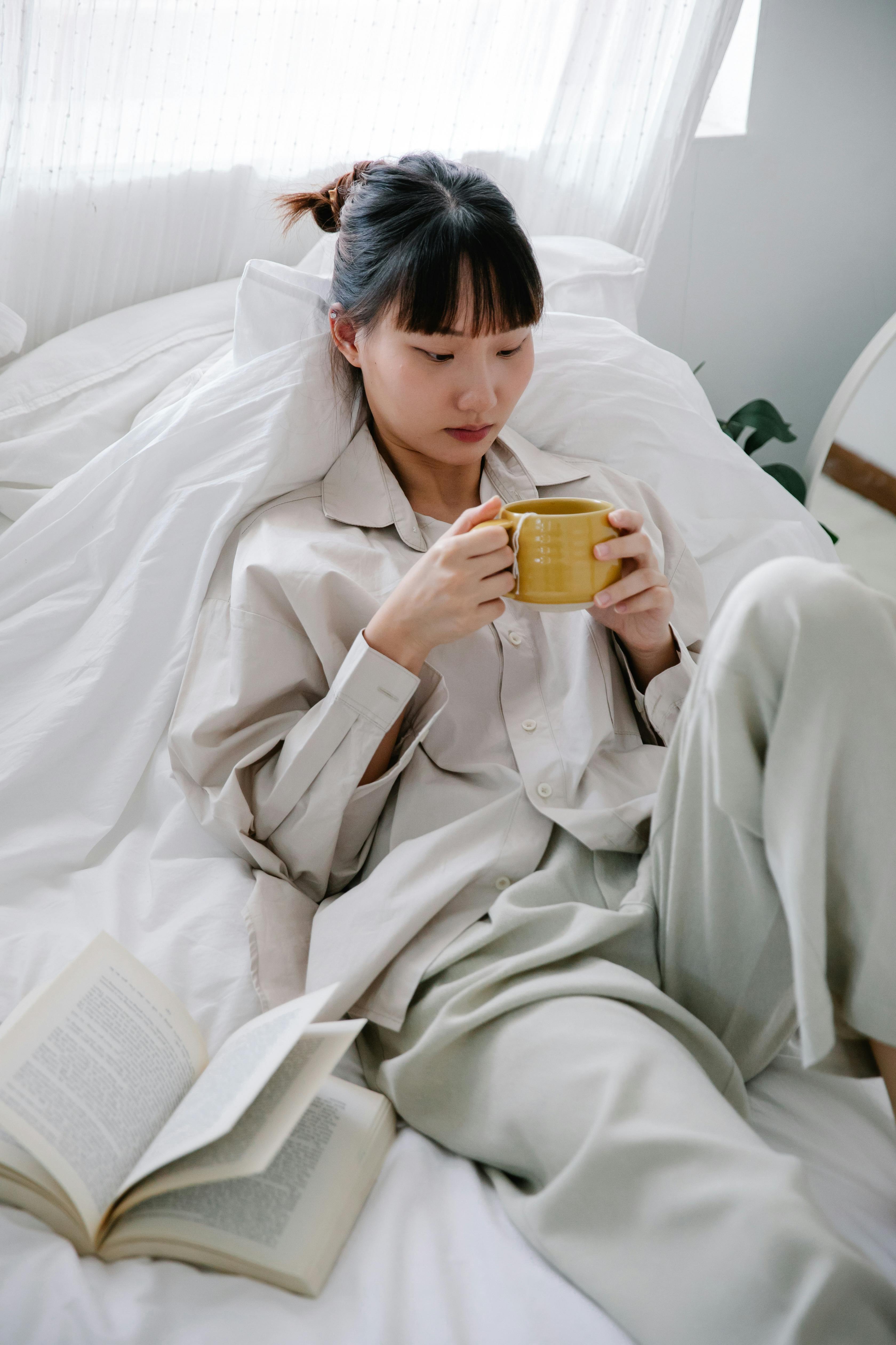 Asian woman enjoying a peaceful morning with coffee and a book in bed.