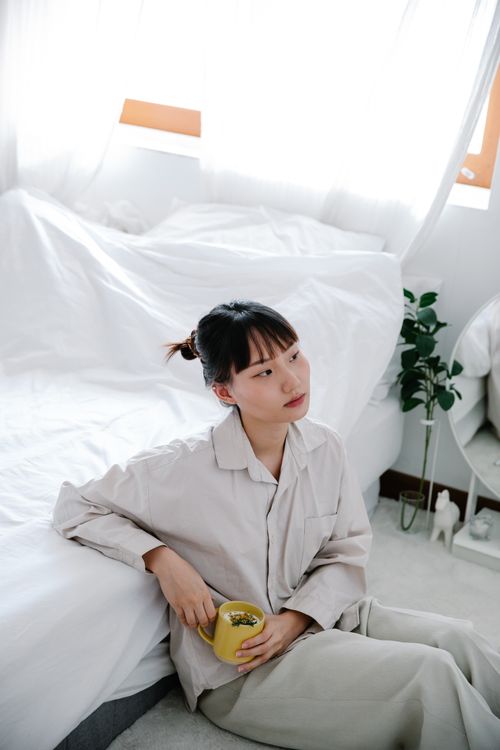Free Asian woman in pajamas sitting on a bed in a bright bedroom, holding a cup. Stock Photo