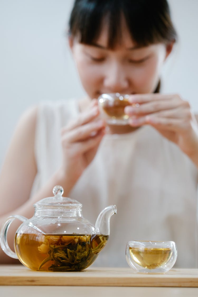 Glass Teapot On A Table And Woman Sipping Tea From A Small Glass