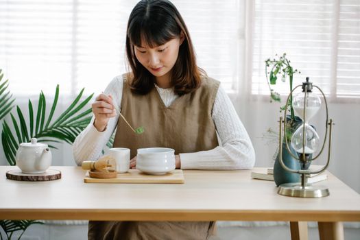 Asian woman preparing matcha tea indoors using a bamboo whisk, emphasizing tradition and calm.