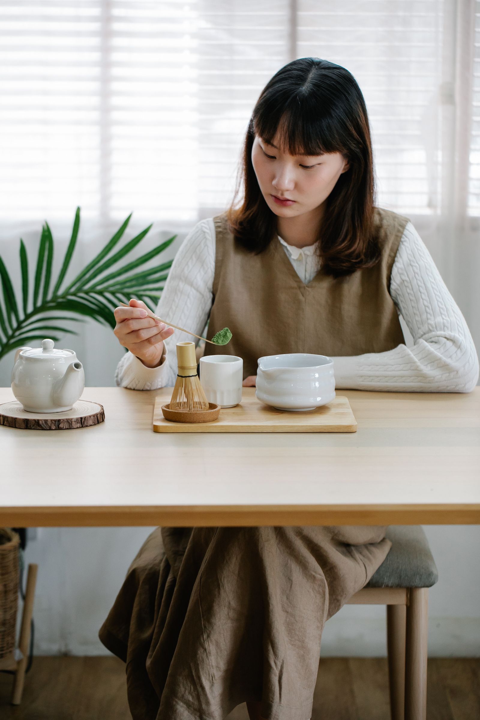 Woman measuring green tea powder beside a bowl, whisk, and teapot at a bright table