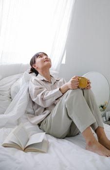 Asian woman in tranquil bedroom, enjoying coffee and a book.